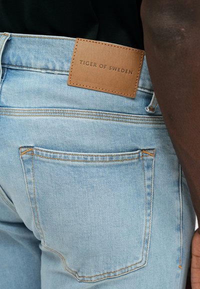 Svetlomodré džíny z denimu s oranžovým prešívaním, so štítkom z kože so značkou "TIGER OF SWEDEN" na zadnom páse.