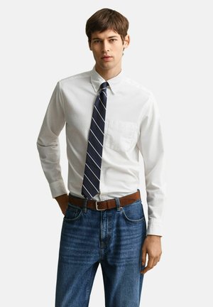 Junger Mann, der ein weißes Hemd mit Knöpfen, eine marineblaue gestreifte Krawatte, einen braunen Gürtel und blaue Jeans trägt, steht mit einer Hand in der Tasche.