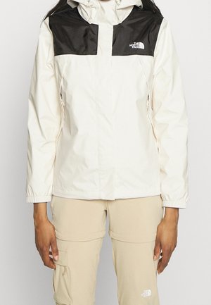 Persona con una chaqueta North Face blanca y negra y pantalones cargo beige, de pie frente a un fondo claro y sencillo.