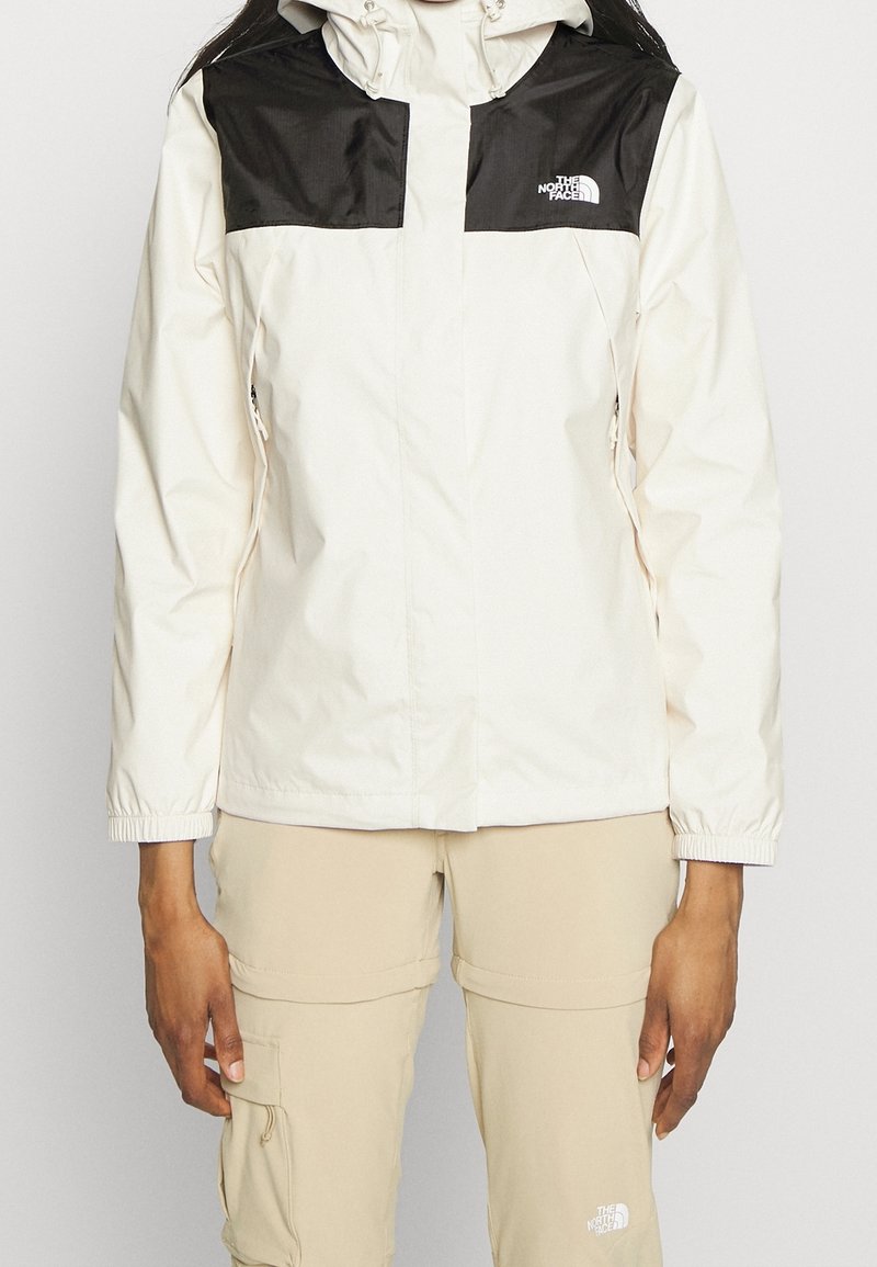 Personne portant une veste North Face blanche et noire avec un pantalon cargo beige, debout devant un fond clair uni.