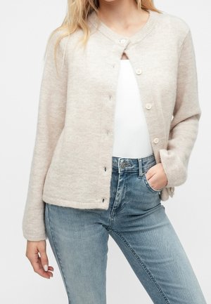 Frau trägt einen hellbeigen, geknöpften Cardigan über einem weißen Top mit einer Hand in der Tasche der hellblauen Jeans vor einfachem Hintergrund.