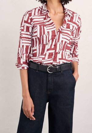 Femme portant une chemise à motifs rouges et blancs avec boutons, manches retroussées, jean bleu foncé et ceinture noire avec boucle argentée.