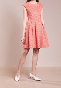 Vestido de coral con estampado floral, mangas de capucha, corsé ajustado y falda acampanada. Combinado con tacones blancos de punta. Textura de tela suave.