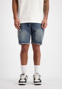 Een persoon staat in een wit t-shirt, versleten blauwe spijkerbroekshorts en zwarte sneakers met witte veters, tegen een effen achtergrond.