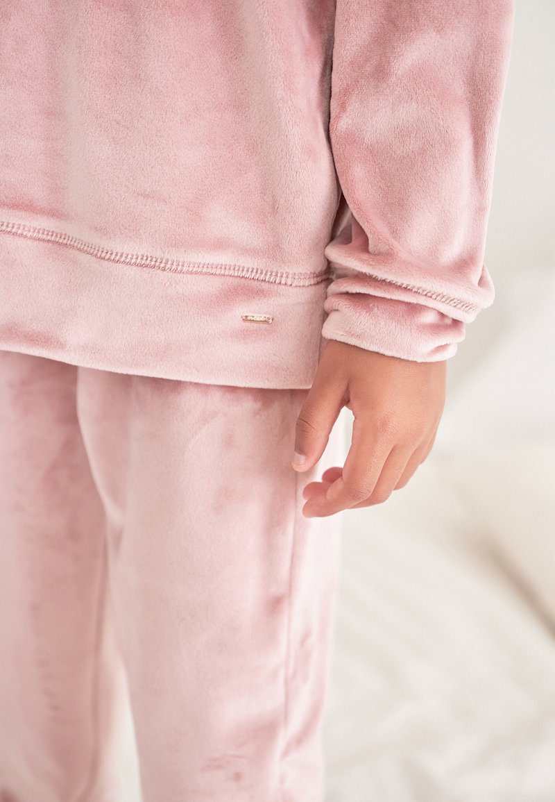 Pink, soft, plush loungewear set featuring a long-sleeve top and matching pants with a small metallic logo detail on the hem.