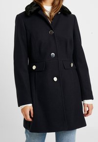 Cappotto in misto lana di un blu scuro con colletto in pelliccia sintetica, bottoni argentati, tasche laterali e design strutturato. Maniche lunghe e fit di lunghezza media.