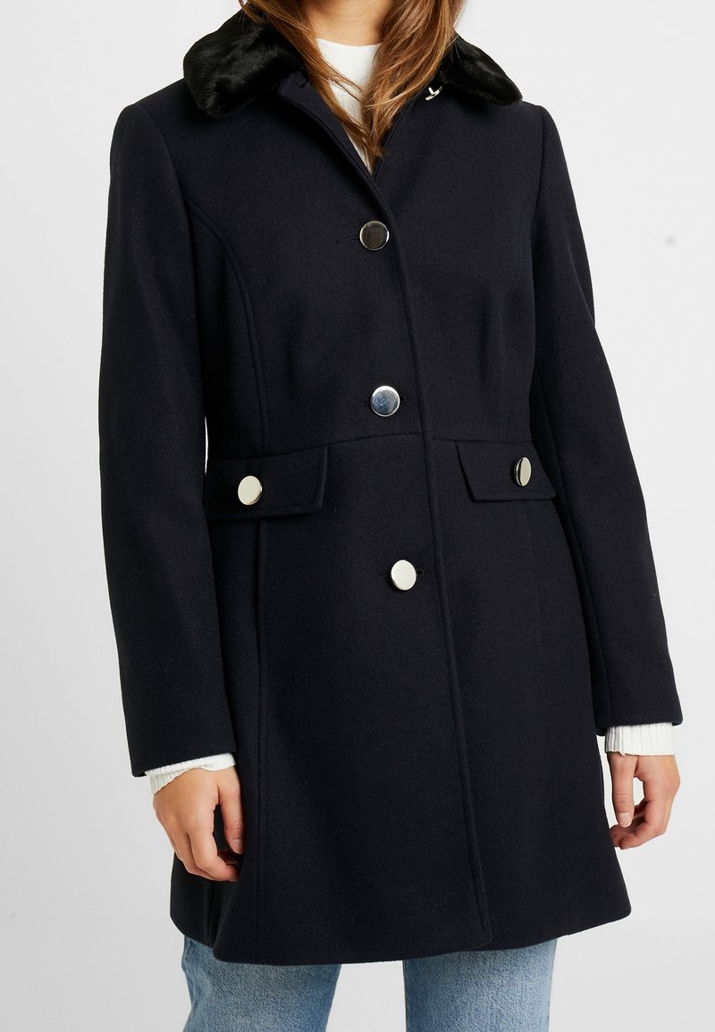 Cappotto in misto lana di un blu scuro con colletto in pelliccia sintetica, bottoni argentati, tasche laterali e design strutturato. Maniche lunghe e fit di lunghezza media.