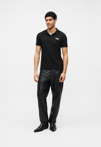 Άνδρας που φοράει μαύρο πόλο με κοντά μανίκια και λογότυπο, μαύρο δερμάτινο παντελόνι και μαύρα μυτερά παπούτσια, στέκεται μπροστά σε λευκό φόντο.