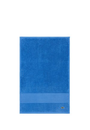Serviette rectangulaire bleue avec une bande texturée près du bord inférieur et un petit logo de crocodile vert dans le coin.