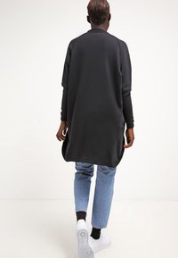 Osoba odcházející, oblečená v dlouhém černém kabátu, světle modrých džínách, černých ponožkách a bílých teniskách na bílém pozadí.