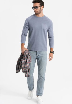 Lange mouwen, lichtblauw T-shirt met een borstzak, gecombineerd met lichtblauwe spijkerbroek en witte sneakers, terwijl hij een geruite jas vasthoudt.