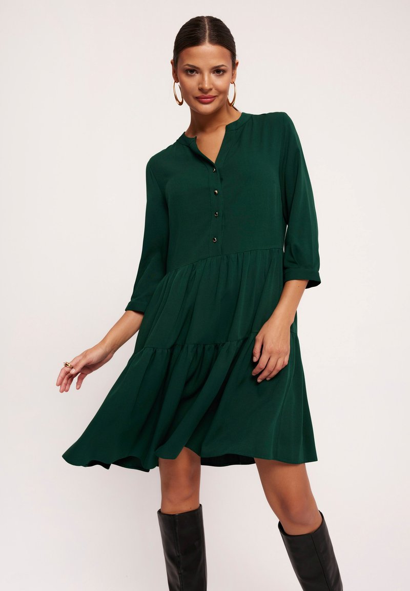 Zielona, warstwowa sukienka z guzikiem przy dekolcie, z rękawami o długości trzech ćwiartek i zwiewnym materiałem, w połączeniu z czarnymi kozakami do kolan. Gładka faktura.