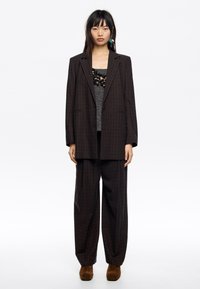 Blazer de cuadros marrón de gran tamaño emparejado con pantalones de pierna ancha a juego. El material parece texturizado; el conjunto presenta una blusa de capas de contraste.