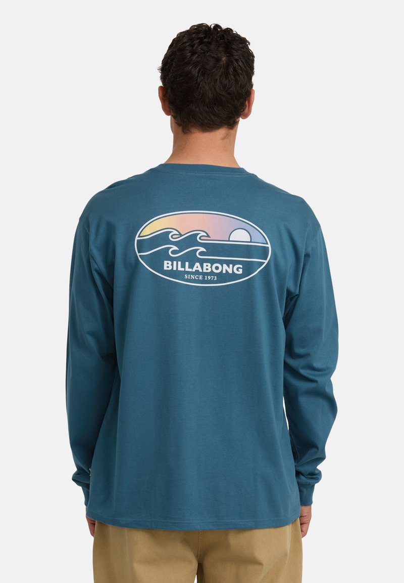 Mężczyzna w turkusowej koszuli z długim rękawem z logo zachodu słońca i fali oraz napisem "BILLABONG SINCE 1973" na plecach, w połączeniu z beżowymi spodniami.