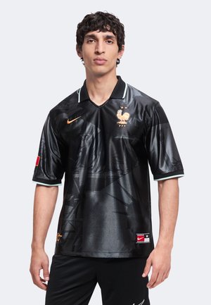 Jeune homme portant un maillot de football noir de la France avec un emblème de coq doré, un logo Nike orange et des motifs graphiques subtils.