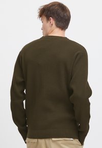 Mørk olivengrønn genser med et teksturert vaffelmønster, lange ermer og ribbestrikket kant, båret av en person sett fra ryggen.
