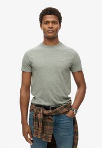Groene gemeleerde t-shirt met korte mouwen. Draag het met blauwe jeans en een geruite shirt om de taille gebonden. Zwarte horloge om de pols.