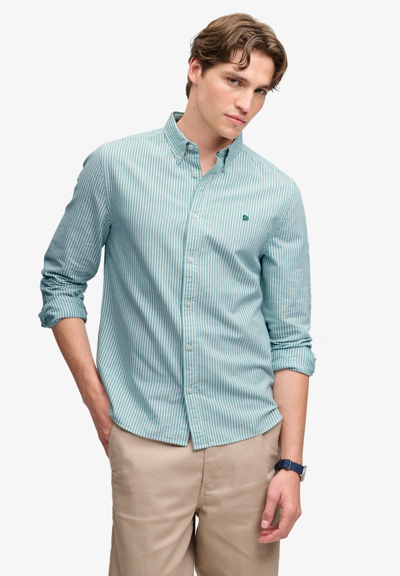 Jeune homme portant une chemise rayée bleu clair à boutons, manches retroussées, et un pantalon beige sur un fond uni.