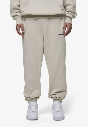 Beige sweatpants with elastic cuffs and waistband. Features black logo on left thigh. Paired with white sneakers. Smooth texture, relaxed fit.
