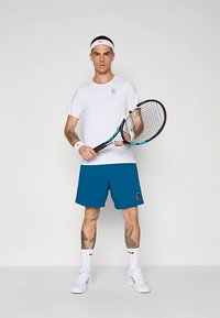 Λευκό πουκάμισο με κοντά μανίκια, μπλε σορτς, κρατώντας ρακέτα τένις. Λευκή κορδέλα στα μαλλιά και βαμβακερά περιβραχιόνια, λευκά παπούτσια αθλητικά. Μονόχρωμα, αθλητικός σχεδιασμός.