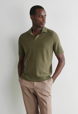 Mężczyzna z krótkimi włosami ubrany w oliwkowo-zieloną, krótkorękawową dzianinową koszulkę polo i beżowe spodnie, stojący z rękami w kieszeniach na tle białej ściany.