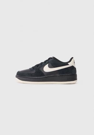 Baskets Nike Air Force 1 noires avec logo swoosh blanc et talon et semelle couleur crème, affichées sur un fond gris clair uni.