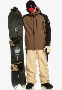 Ein Snowboarder hält ein schwarzes Snowboard mit braunen und cremefarbenen Bindungen. Er trägt eine braun-schwarze Jacke, cremefarbene Hose und schwarze Stiefel. Rote Goggles.