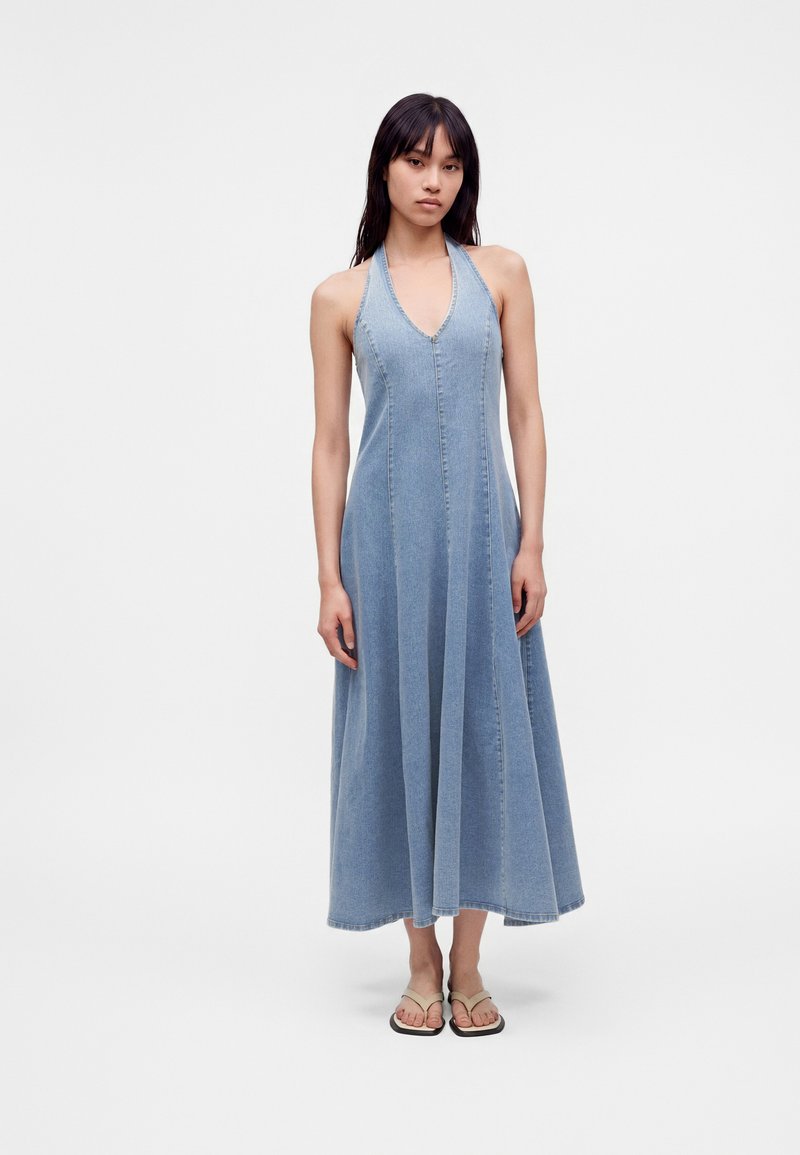 Γυναίκα που φοράει μπλε denim αμάνικο φόρεμα halter με βαθύ V ντεκολτέ και μπεζ σανδάλια, στέκεται μπροστά σε λευκό φόντο.