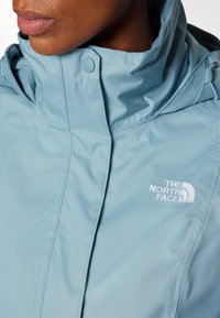 Šviesiai mėlyna striukė su aukštu apykakle, su balta "The North Face" logotipu, lygaus paviršiaus ir snapeliu uždarymo detale.