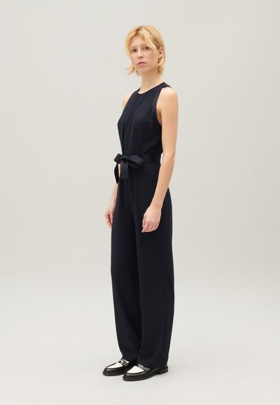 Combinaison sans manches bleu marine avec un corsage ajusté et un pantalon large, agrémentée d'une ceinture nœud assortie. Présente une poche latérale unique et une texture lisse.
