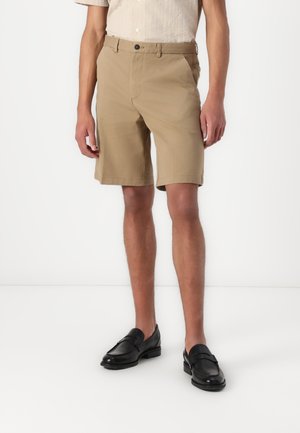 Shorts pour hommes beiges à coupe droite, avec poches avant et fermeture à un seul bouton. Portés avec des mocassins noirs. Tissu en coton lisse.