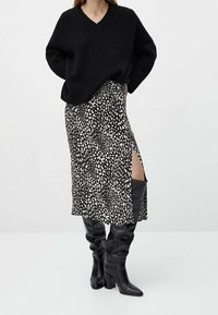 Pull en maille noir oversize avec un col en V, associé à une jupe mi-longue arborant un motif abstrait blanc et une fente haute, port é avec des bottes noires montant jusqu'aux genoux.