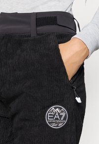 Μαύρο παντελόνι κορντούρας με υφασμάτινη επιφάνεια, με μαύρη ζώνη στη μέση και λογότυπο μπροστά. Περιλαμβάνει λεπτομέρεια με τσέπη με φερμουάρ.