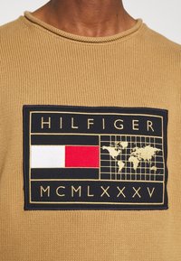 Beiger Strickpullover mit einem rechteckigen marineblauen Patch, auf dem "HILFIGER", eine Weltkarte und rot, weiß und rot gefärbte Blöcke zu sehen sind.