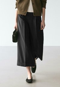 Schwarze Hosen mit weitem Bein aus weichem Stoff, mit umgeschlagenem Saum und glattem Finish, kombiniert mit einer braunen, gerippten Strickjacke und schwarzen Ballerinas.