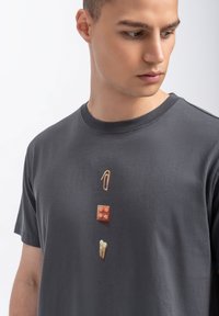 Grijze katoenen t-shirt met een grafisch ontwerp met een paperclip, een bruine blok en een tandillustratie, verticaal op de voorkant gerangschikt.