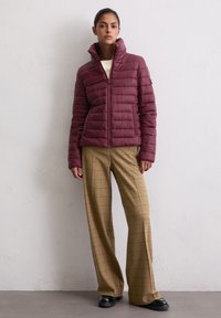 Bordeaux pufferjack met een hoge kraag, ritssluiting aan de voorkant en een gewatteerd ontwerp, gecombineerd met beige geruite brede broek en zwarte schoenen.