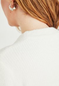 Κοντινό πλάνο από τον λαιμό και το αριστερό αυτί ενός ατόμου με κόκκινα μαλλιά, που φοράει ένα ριμπ (ραμμένο) λευκό πουλόβερ και ένα ασημένιο σκουλαρίκι.