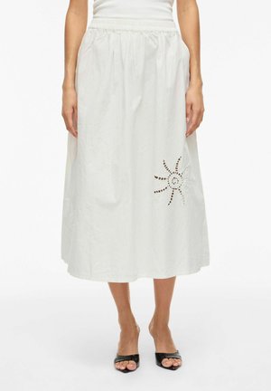 Femme portant une jupe midi blanche avec une taille froncée et un motif découpé en forme de soleil sur le côté droit, associée à des talons noirs à bouts ouverts.