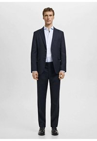 Temno moder oblečen kostim s suknjičem z enim gumbom in hlačami z ravnim sprednjim delom, kombiniran z svetlo modro srajco in črnimi čevlji.