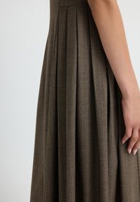 Vestido marrón con pliegues, hecho de una tela texturada, que presenta un cuerpo ajustado y una falda fluida, con detalles de patrón sutiles. Mano visible en el lateral.