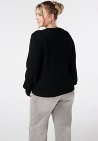 Zwarte gebreide trui met een ronde hals, lange mouwen en een geribde zoom. Gecombineerd met lichtgrijze wijde broekspijpen, gezien vanaf de achterkant.