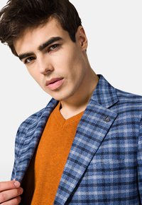 Niebieska marynarka w kratę z teksturowanym wykończeniem, z czarnym akcentem. Pod spodem widoczny jest pomarańczowy sweter z dekoltem w serek.