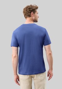 Niebieska bawełniana koszulka z krótkim rękawem i okrągłym dekoltem, charakteryzująca się gładką fakturą i minimalistycznym wzorem z tyłu, noszona z jasnobeżowymi spodniami.