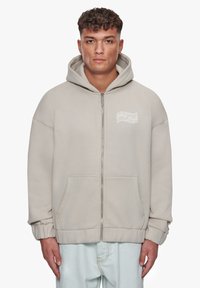 Beige zip-op sweatshirt med stor hætte, frontlommer og hvidt trykt logo på venstre bryst. Lavet af blødt, struktureret stof med manchetter ved ærmerne.