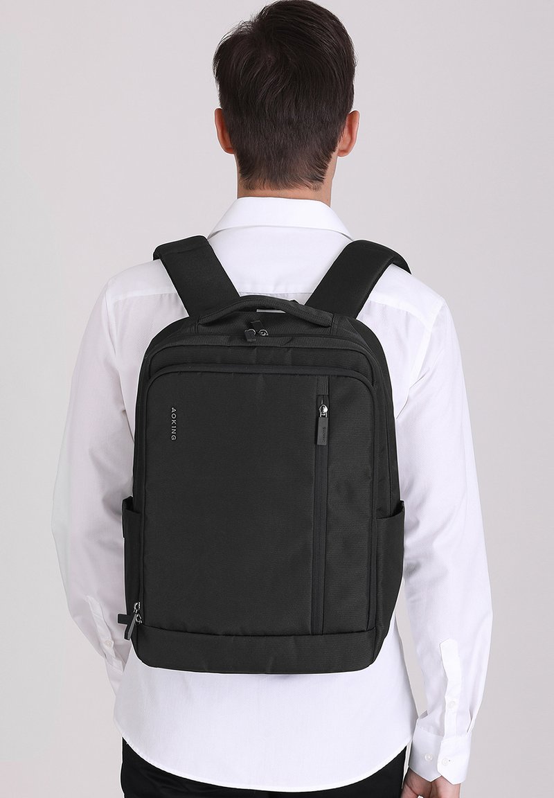 Sac à dos en tissu noir avec des bretelles rembourrées, une poche zippée à l'avant et une poche latérale en mesh. Design épuré, forme rectangulaire et style minimaliste.