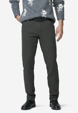 Mann trägt dunkelgraue Hose, schwarzen Gürtel, schwarze Schuhe und einen grauen Pullover mit weißen Mustern, steht mit einer Hand in der Tasche.