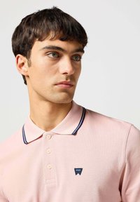 Jasnoróżowa koszulka polo z teksturowanej tkaniny, z granatowym prążkowanym kołnierzykiem, dwoma guzikami i małym niebieskim logo po lewej stronie.