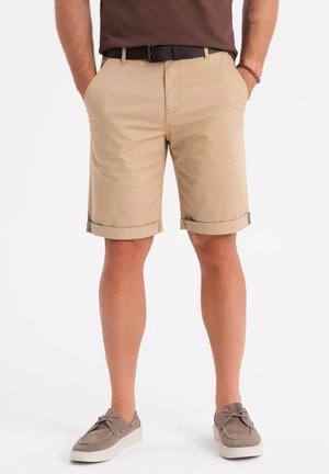 Beige Baumwoll-Chino-Shorts mit umgeschlagenen Säumen, zwei vorderen Taschen und einem versteckten Rückentaschen-Detail; kombiniert mit grauen Wildlederschuhen.