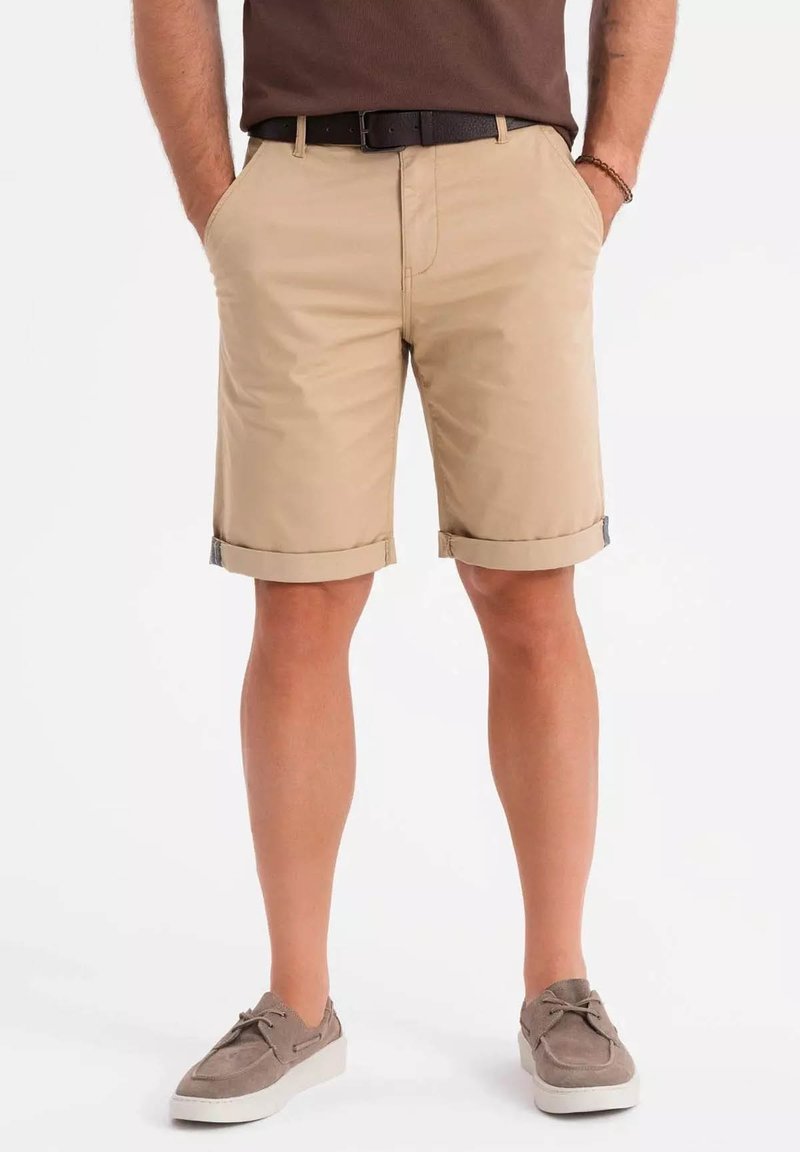Shorts en coton beige avec ourlets retournés, deux poches avant et une poche arrière cachée ; associés à des chaussures en daim grises.
